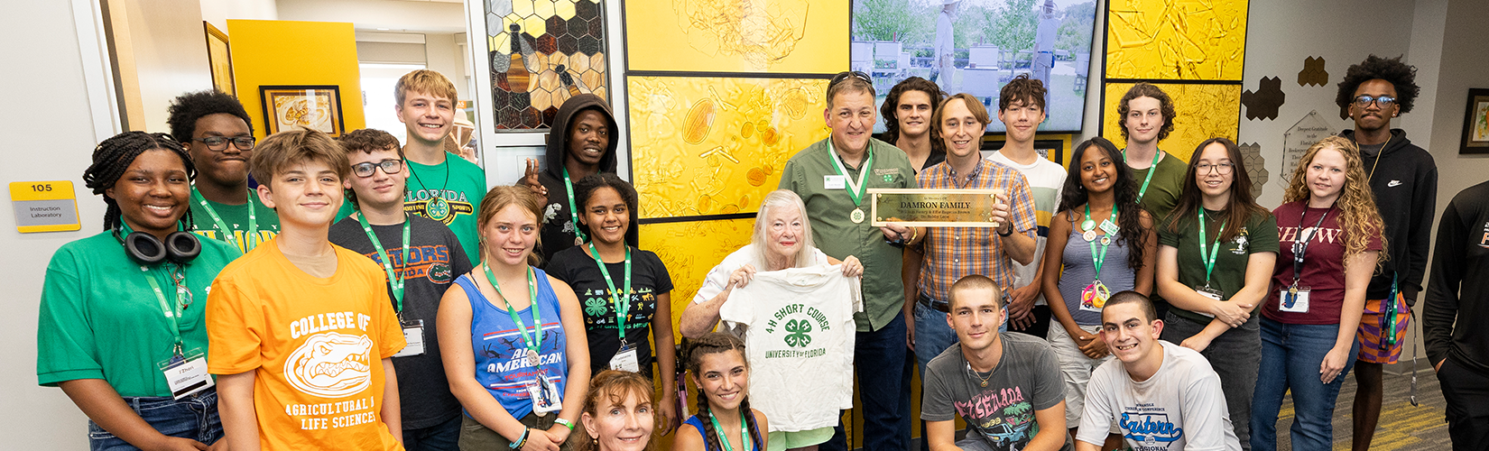 4-H University students, Vera Damron (holding a white 4-H Short Course t-shirt), Chris Oster (holding the Damron Family Office sign) and Todd Bundy (wearing 4-H Hall of Fame medallion) pose in the UF/IFAS Honey Bee Research & Extension Laboratory lobby.