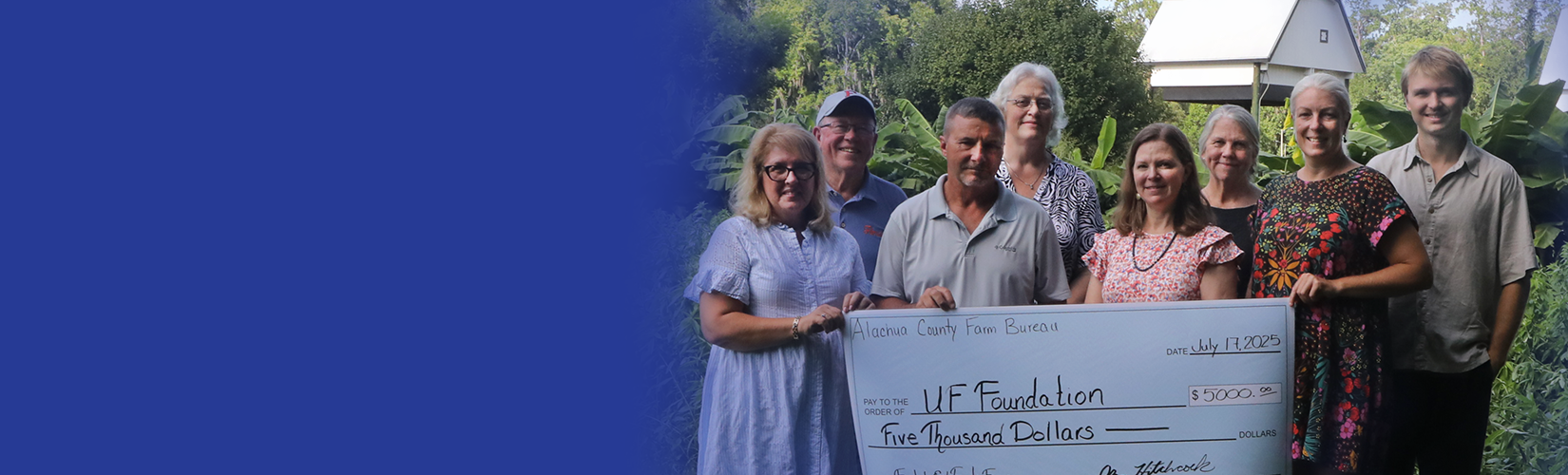 A group of people holding a giant $5,000 check outside the UF Field & fork farm and Gardens. A white UF bat house is visible among the trees in the distance.